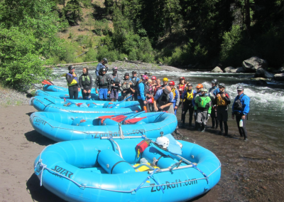 Large group of whitewater guide school students on a river bank awaiting instructions.